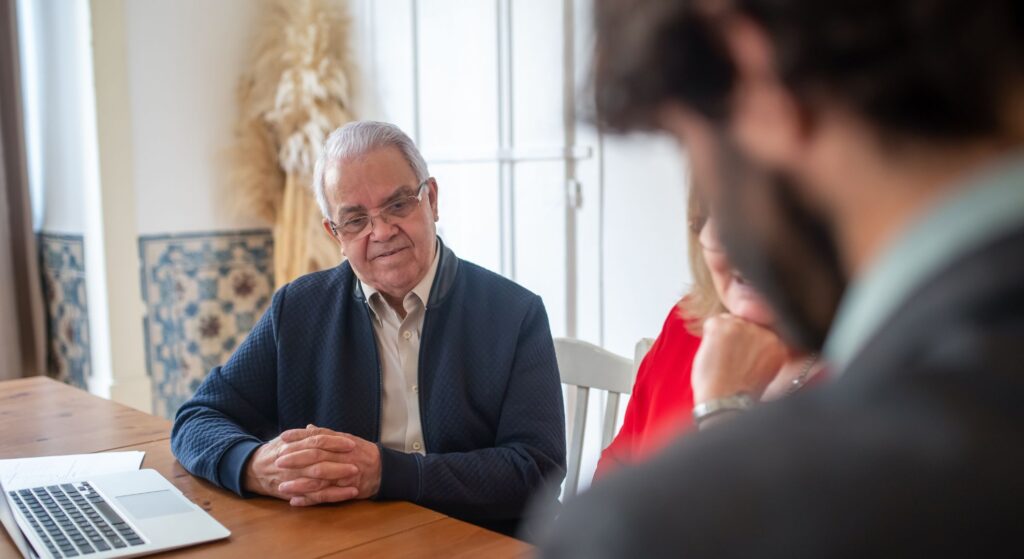 mature man sits at table with laptop while turned to look at person in foreground